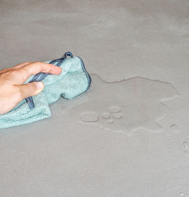 Eine Hand wischt mit einem türkisfarbenen Mikrofasertuch Wasser von einer glatten, grauen Oberfläche auf.