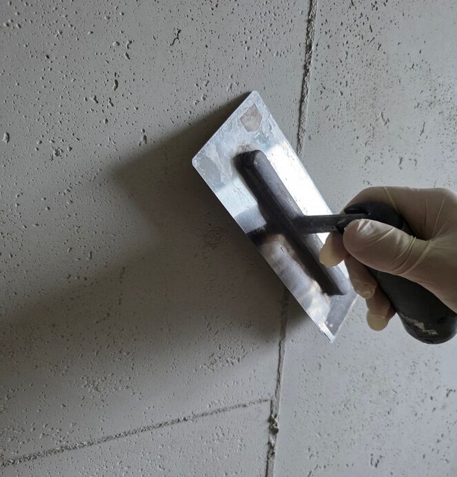 Eine Person mit weißen Handschuhen glättet mit einem Spachtel eine raue Betonwand mit Spachtelmasse, um eine glatte Oberfläche zu erzeugen.